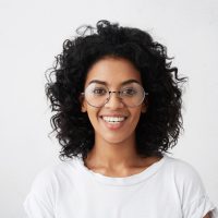 Portrait of positive carefree dark-skinned girl dressed casually smiling broadly, posing isolated at studio wall, having happy joyful look, relaxing indoors. Human facial expressions and emotions